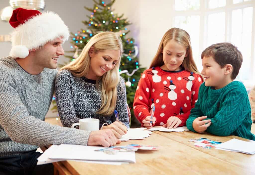 A family sits at a table, writing cards together. A Christmas tree decorated with lights is in the background. The father wears a Santa hat, while the children wear festive sweaters.