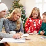 A family sits at a table, writing cards together. A Christmas tree decorated with lights is in the background. The father wears a Santa hat, while the children wear festive sweaters.