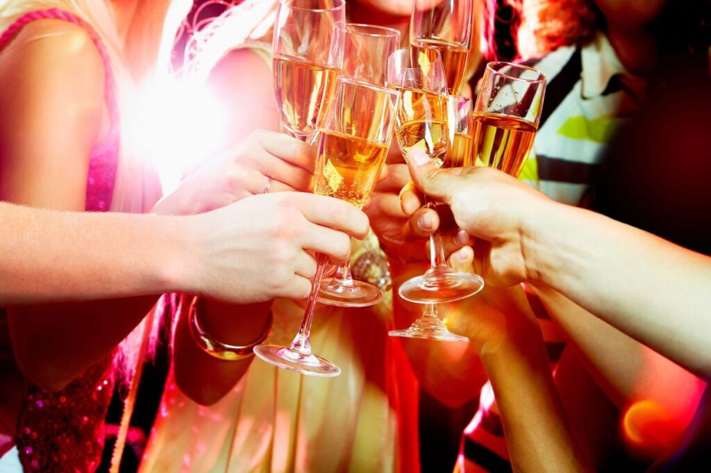 A group of people clink glasses filled with champagne in a celebratory New Year’s Eve toast, with bright lights in the background.