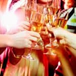 A group of people clink glasses filled with champagne in a celebratory New Year’s Eve toast, with bright lights in the background.