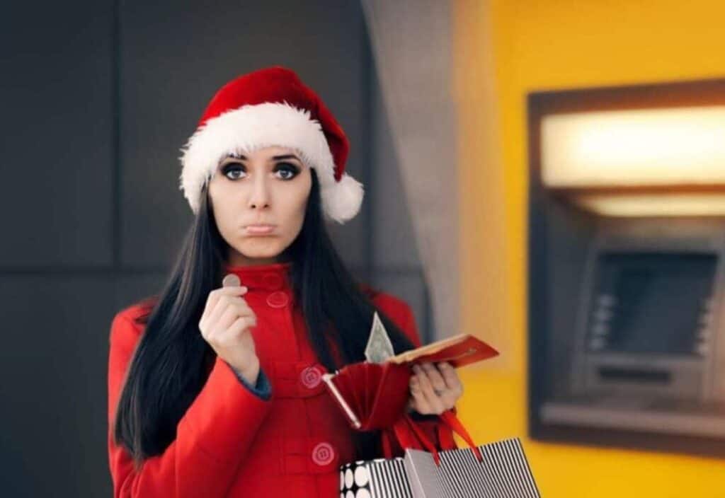 A woman in a Santa hat and red coat stands in front of an ATM, holding a coin and an empty wallet, looking concerned about her holiday shopping.