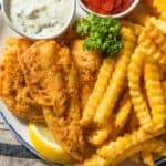 A plate of crispy fried fish and crinkle-cut fries with ketchup and tartar sauce on the side, garnished with parsley and a lemon slice.