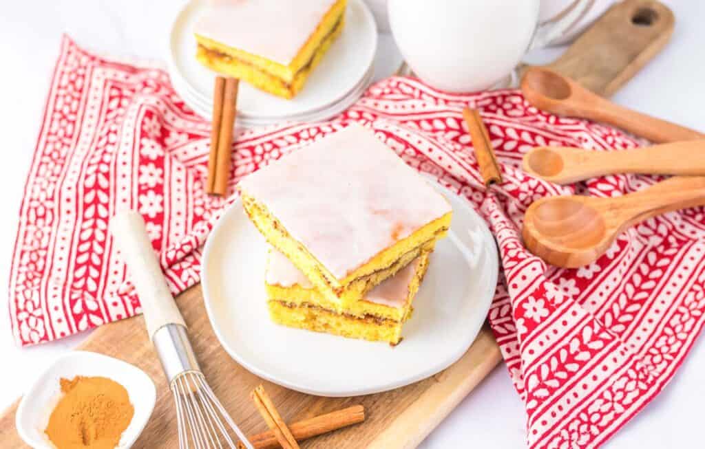 Two slices of frosted cake on a white plate, surrounded by cinnamon sticks and measuring spoons on a red patterned cloth.