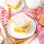 Two slices of frosted cake on a white plate, surrounded by cinnamon sticks and measuring spoons on a red patterned cloth.