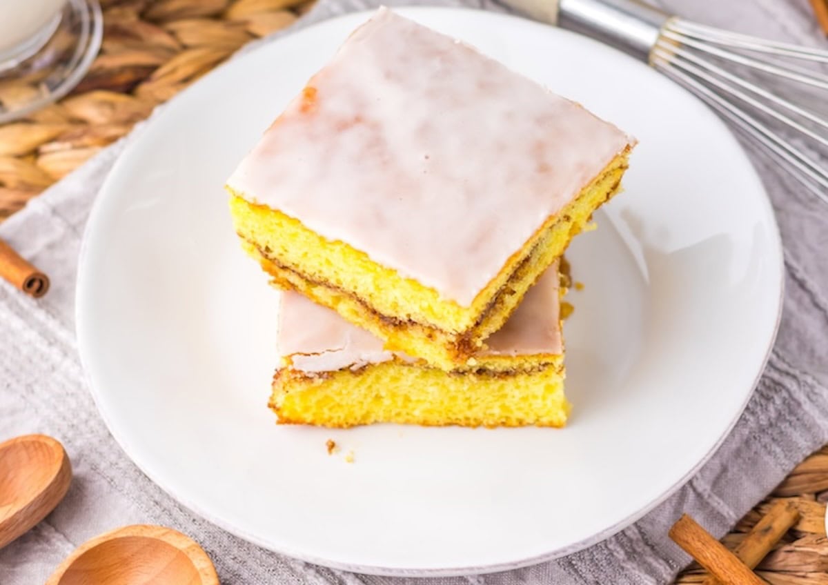 Two slices of frosted yellow cake are stacked on a white plate, placed on a textured tablecloth. A whisk and wooden utensils are visible in the background.