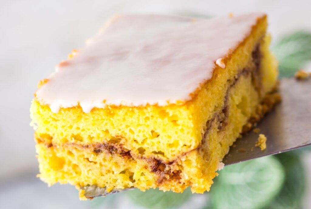 A slice of yellow cake with cinnamon swirls and a sugar glaze on top, served on a spatula.