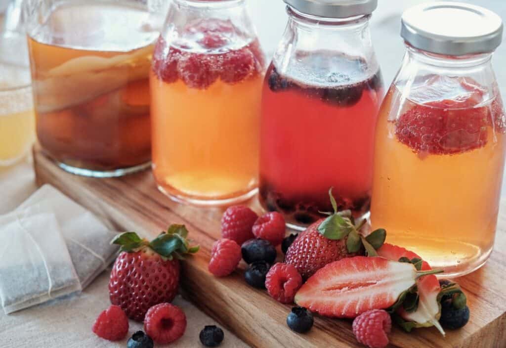 Four glass bottles of fruit-infused drinks on a wooden board, surrounded by fresh strawberries, raspberries, and blueberries.