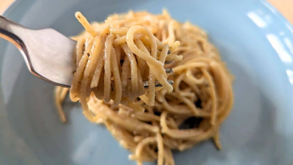 Close-up of a fork holding a serving of spaghetti with black pepper, resting above a plate of more spaghetti in a light blue dish.