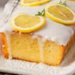 A glazed lemon loaf cake topped with lemon slices and sprigs of rosemary on a white rectangular plate.