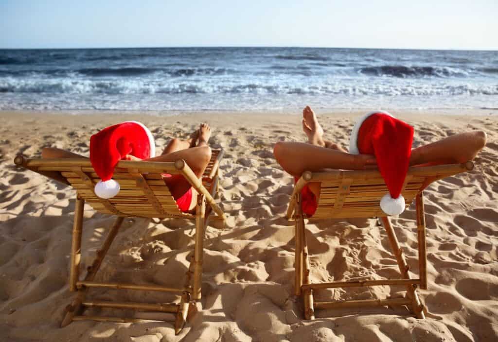 Two people wearing Santa hats relax on wooden beach chairs facing the ocean on a sandy beach.