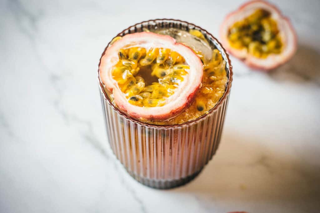 A glass filled with a dark beverage topped with a halved passion fruit, on a light marble surface. Another half of the passion fruit is in the background.