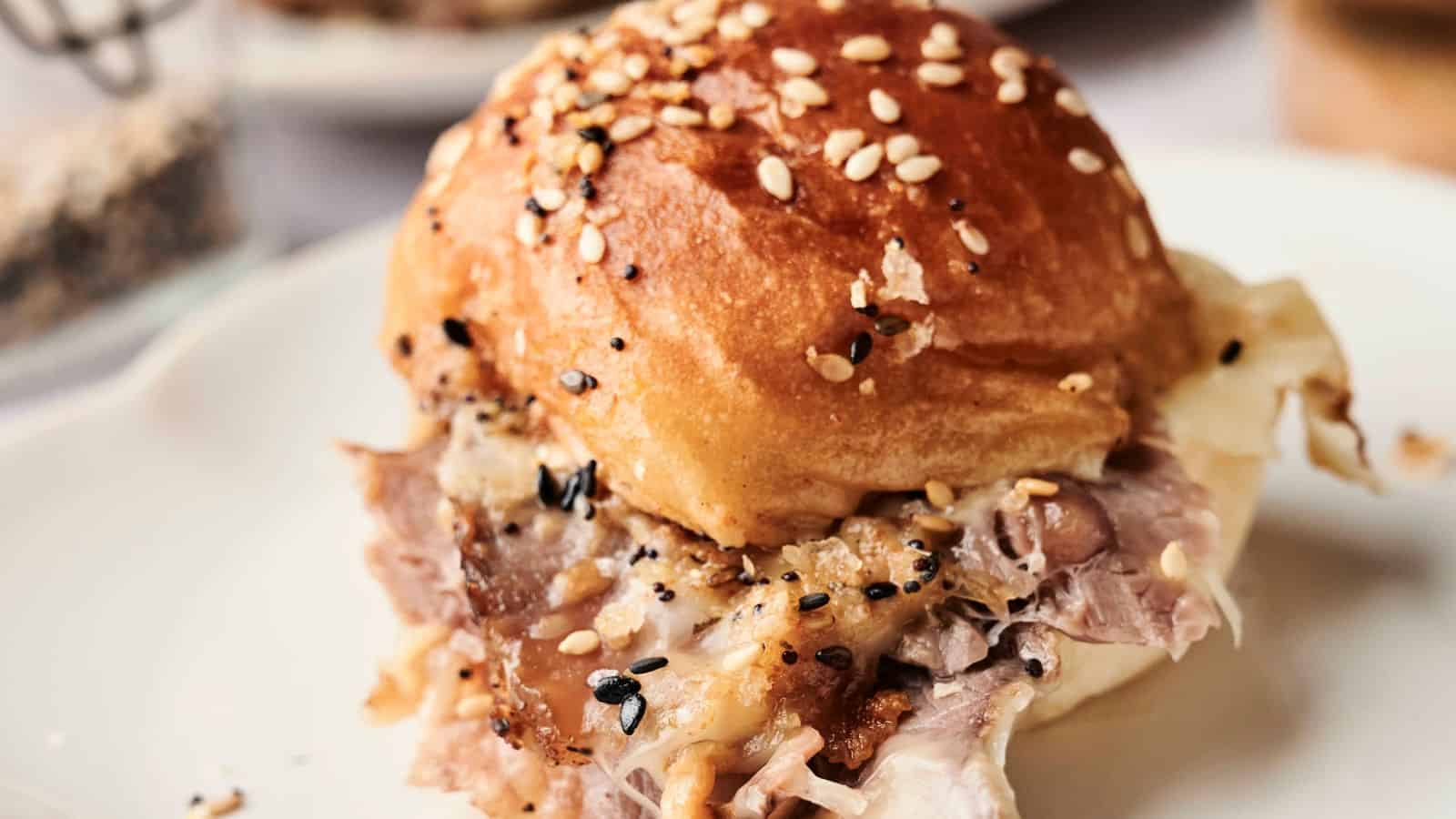 A sandwich with shredded meat and melted cheese on a sesame seed bun is placed on a white plate.