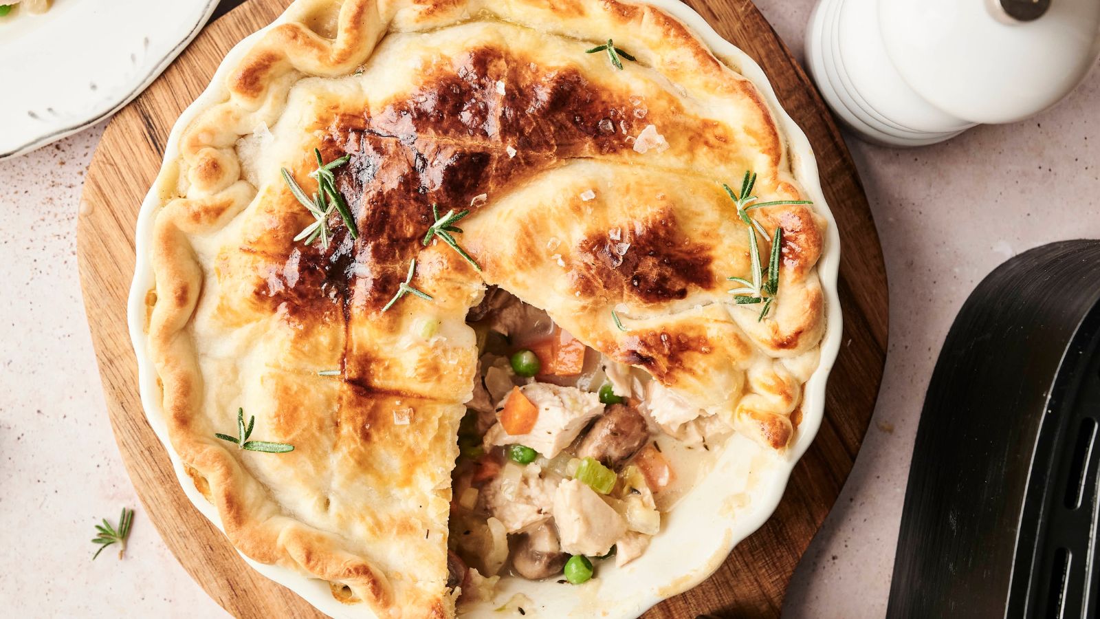 A freshly baked chicken pot pie with a slice missing, revealing chunks of chicken, peas, and carrots inside. Garnished with rosemary on a wooden board.