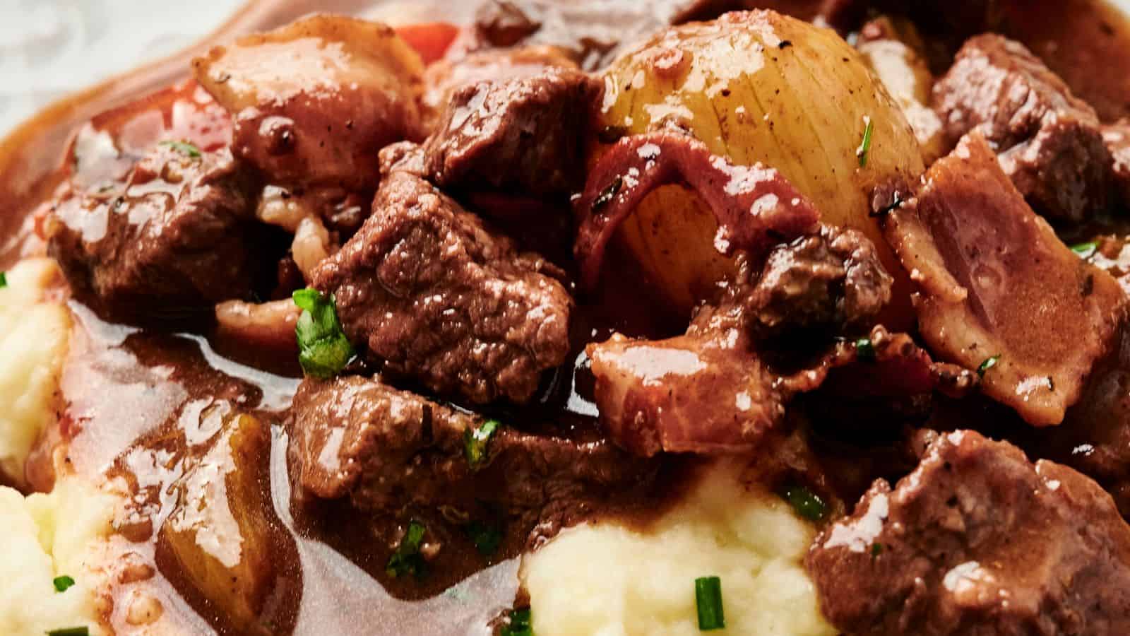 Close-up of a savory beef stew with chunks of beef, bacon, onion, and vegetables in a rich, glossy sauce, served over creamy mashed potatoes.