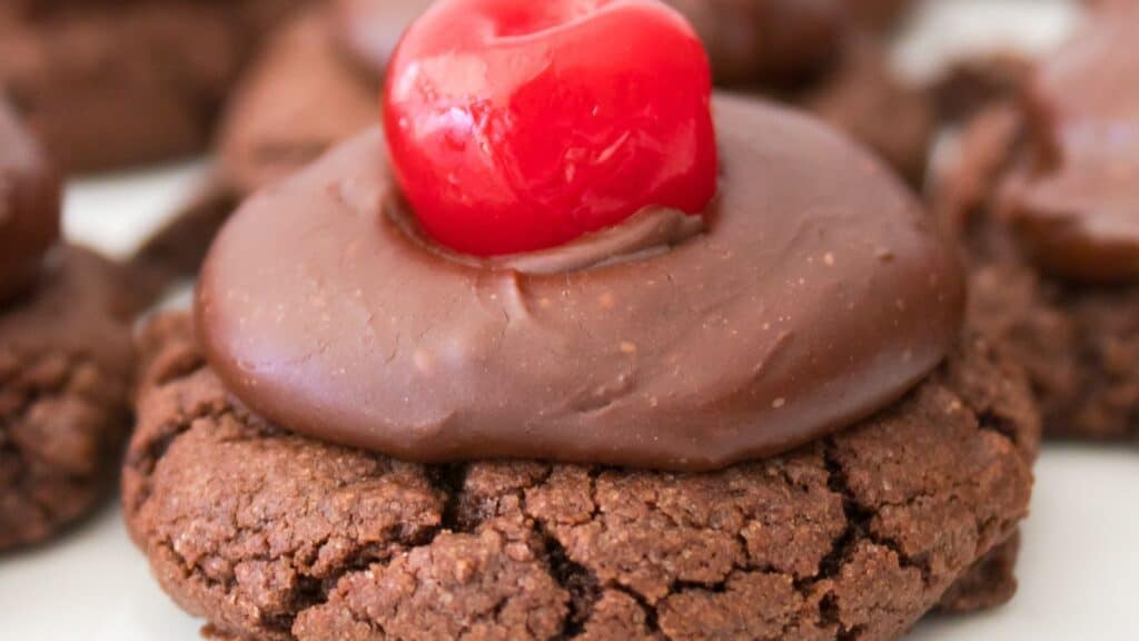 A chocolate cookie topped with glossy chocolate frosting and a bright red cherry.