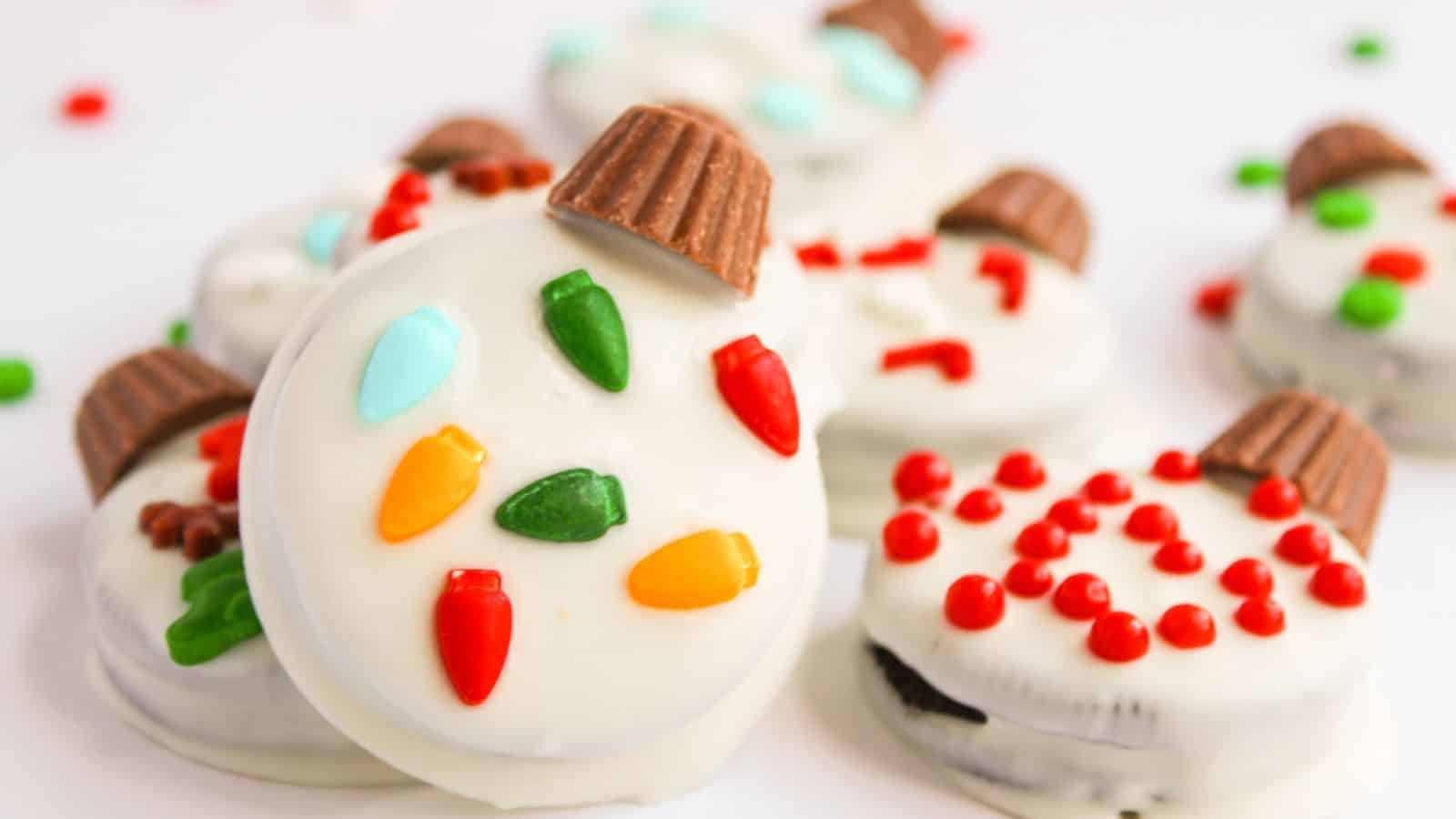 A close-up of Oreo Christmas Ornament Cookies, elegantly coated in white chocolate and adorned with colorful candy lights, red sprinkles, and chocolate pieces on top.