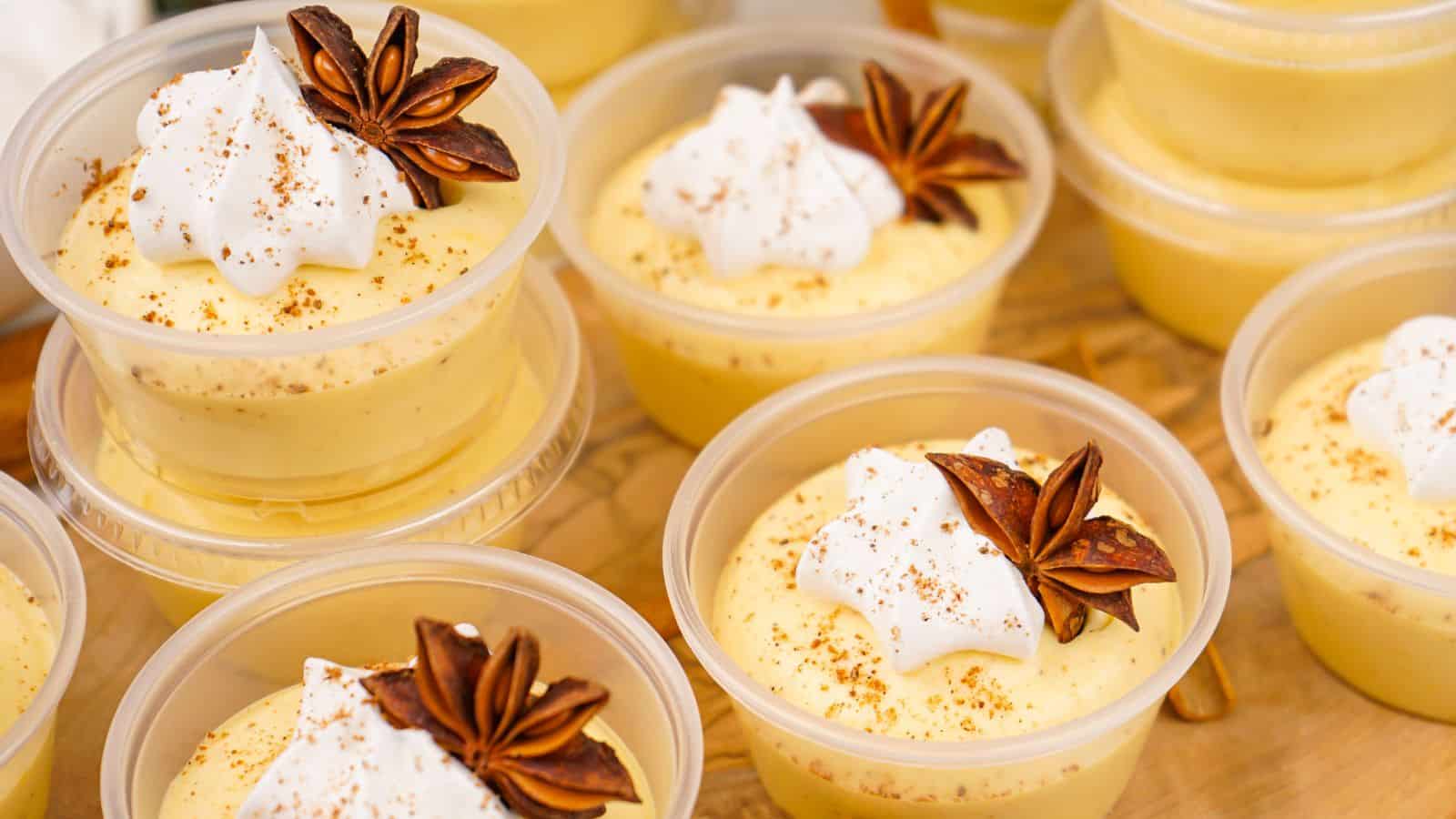 Plastic cups filled with creamy yellow eggnog pudding shots are topped with whipped cream, star anise, and a sprinkle of cinnamon.