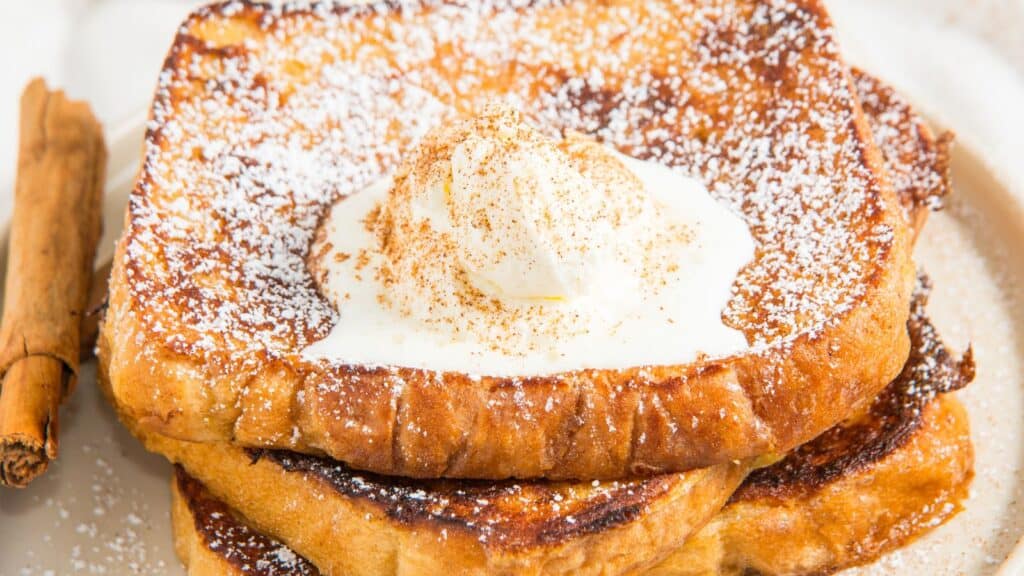 A stack of French toast topped with whipped cream and dusted with powdered sugar, next to a cinnamon stick on a plate.