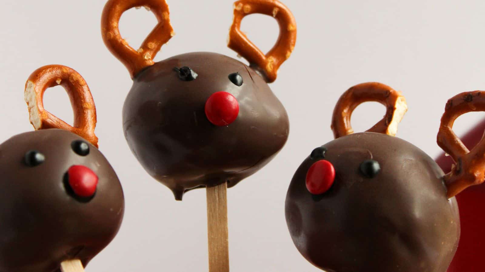 Three chocolate cake pops shaped like reindeer, with pretzel antlers and red candy noses, on wooden sticks.