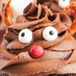 Chocolate cupcake with swirled frosting decorated to resemble a face with candy eyes, a red candy nose, and pretzel antlers.