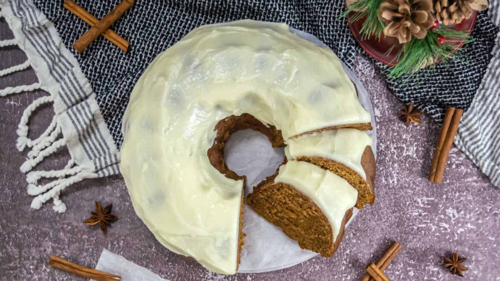 bundt cake with slices cut, on a dark cloth, surrounded by cinnamon sticks and star anise. A small decorative pine arrangement is nearby.