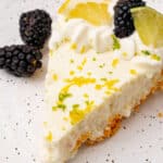 A slice of pie topped with whipped cream, blackberries, a lemon wedge, and a lime wedge, garnished with lemon and lime zest on a speckled plate with a fork.