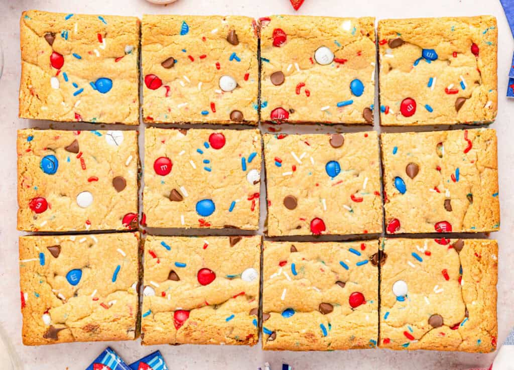 Twelve colorful cookie bars with red, white, and blue candies and sprinkles, arranged in a grid pattern on a light surface.