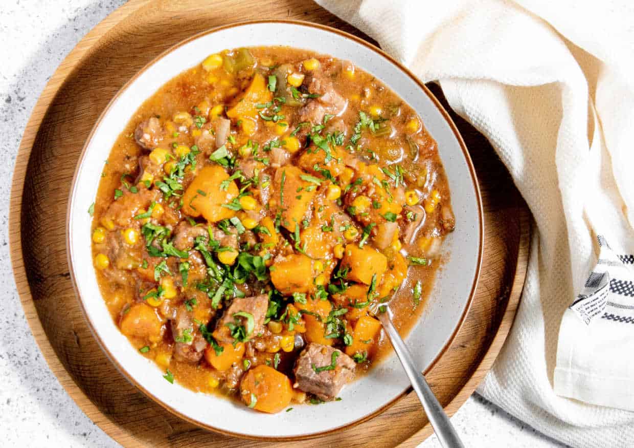 A bowl of beef stew with chunks of beef, carrots, potatoes, and corn, garnished with herbs. A spoon rests in the bowl, and a cloth napkin is nearby on a wooden plate.