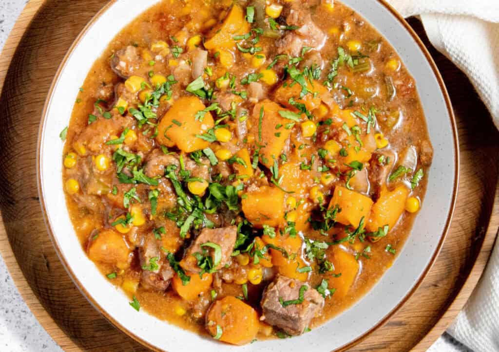 A bowl of stew with beef, corn, and cubed vegetables, garnished with chopped herbs.