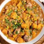 A bowl of stew with beef, corn, and cubed vegetables, garnished with chopped herbs.