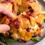 Hand reaching for a piece of baked pull-apart bread with visible apple chunks and cranberries, garnished with rosemary on a gray plate.