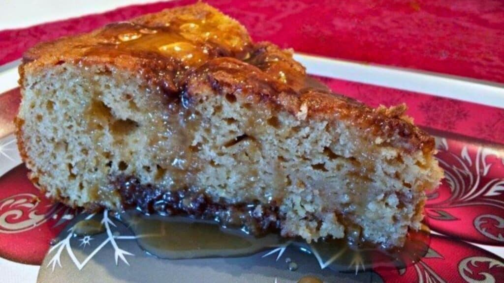 A slice of moist caramel cake on a decorative red and silver plate, with caramel sauce drizzling onto the surface.