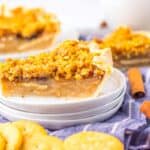 Slice of pie with crumb topping on a stack of plates, surrounded by crackers and cinnamon sticks.