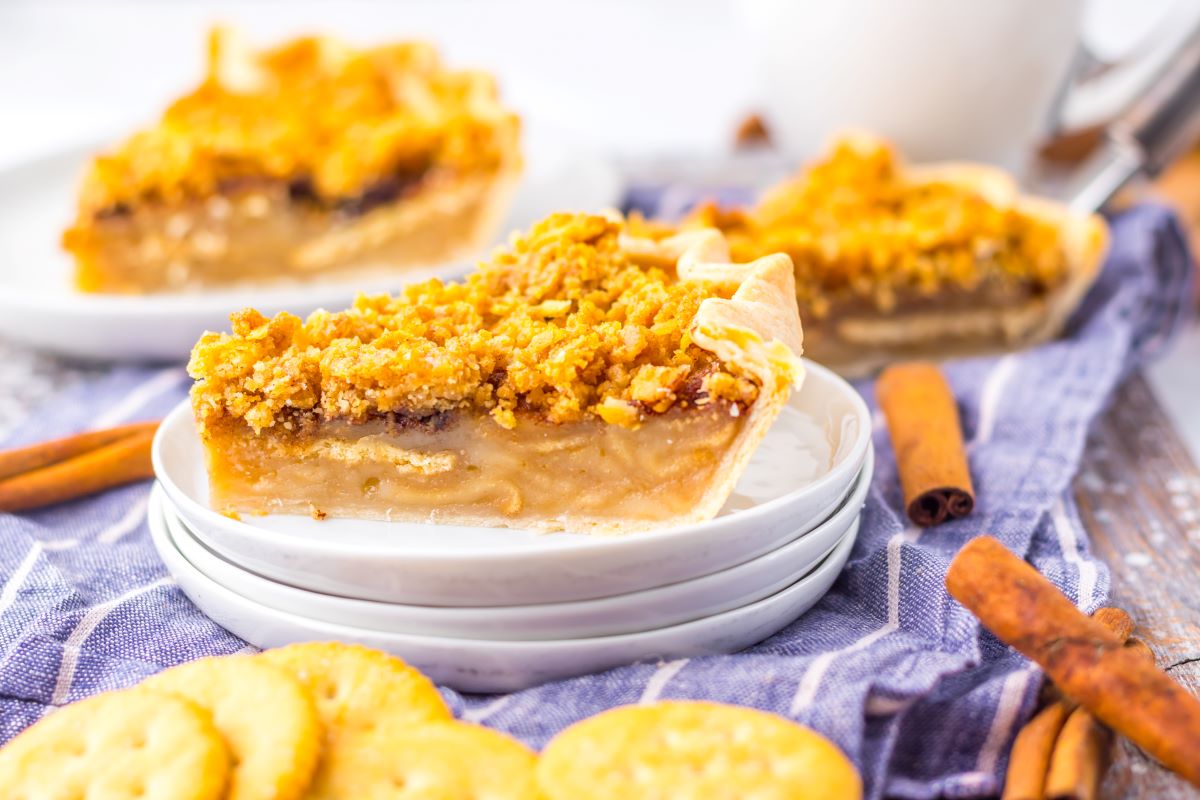 Slice of pie with crumb topping on a stack of plates, surrounded by crackers and cinnamon sticks.