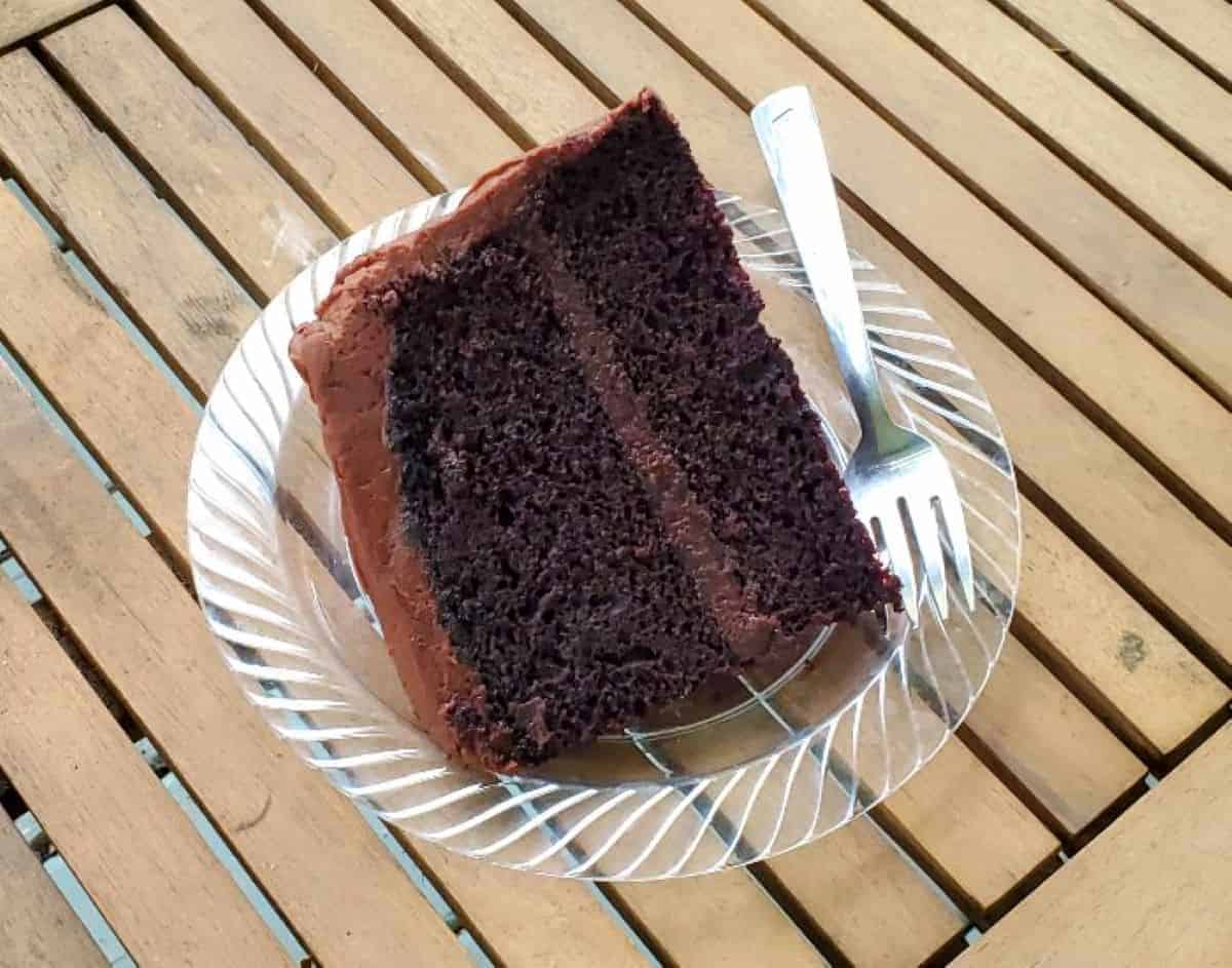 Image shows a slice of chocolate frosted chocolate cake on a plate sitting on a wooden table.