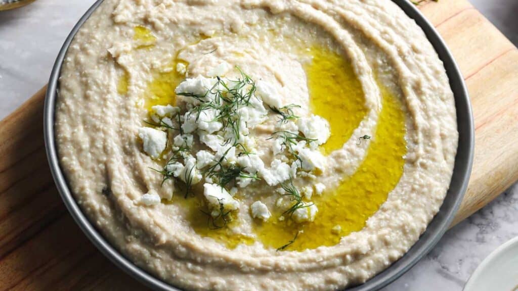 A bowl of hummus topped with crumbled feta cheese, olive oil, and fresh dill on a wooden cutting board.