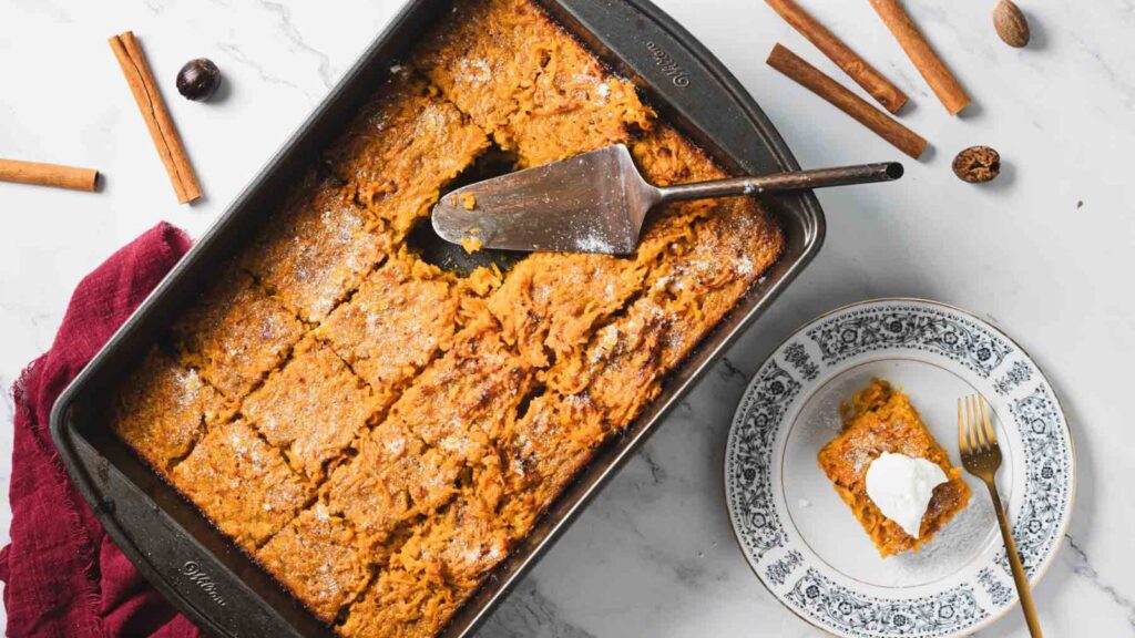 A tray of baked pumpkin dessert cut into squares, with a serving spatula. A piece on a plate with a dollop of cream next to a fork. Cinnamon sticks and nutmeg are scattered around.