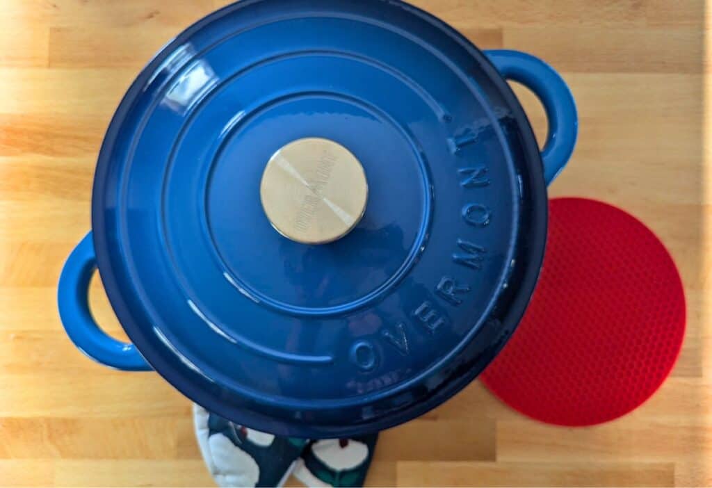 A blue enameled cast iron Dutch oven on a wooden surface, next to a red silicone trivet and a pair of oven mitts.