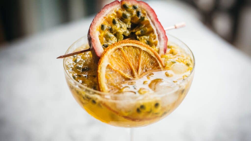 A cocktail glass filled with a yellow drink, topped with half a passion fruit, a slice of orange, and ice cubes.