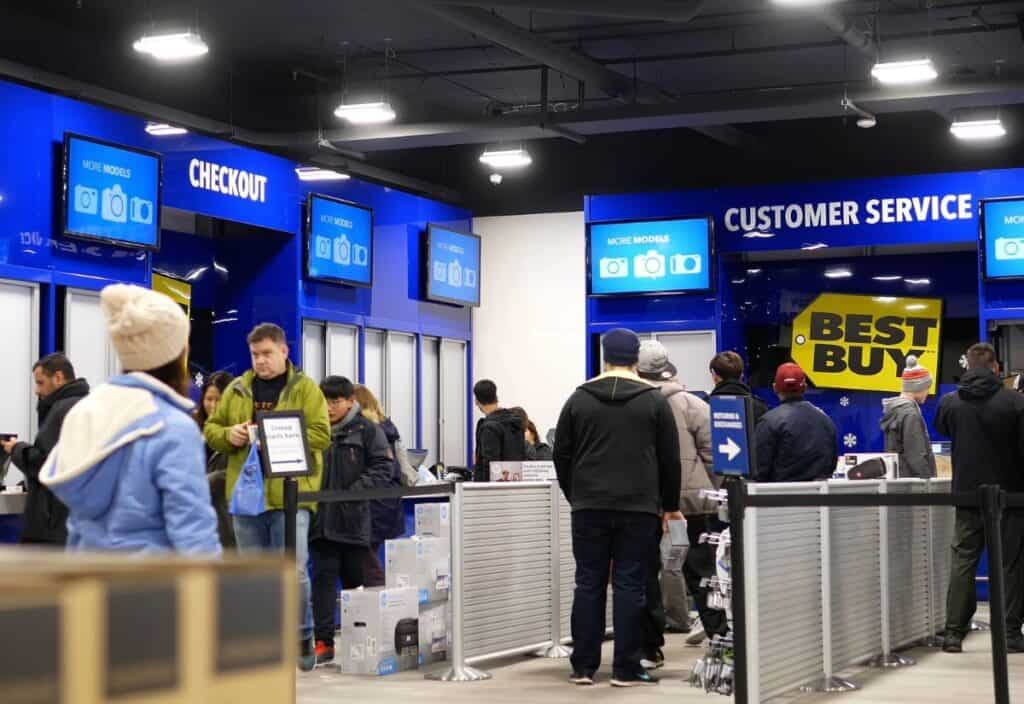 People wait in line at a Best Buy customer service and checkout area, juggling holiday returns. Bright blue signs and electronic displays are visible, adding to the buzzing post-festive atmosphere.