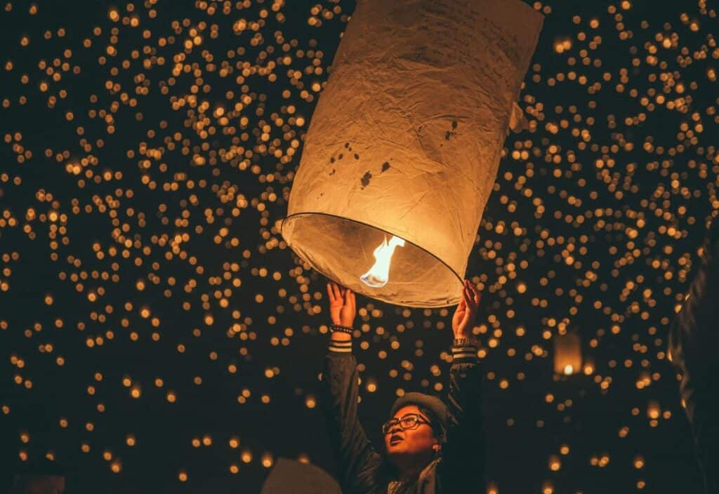 Person releasing a large paper lantern into a night sky filled with numerous glowing lanterns.