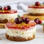 Mini cheesecakes topped with red jelly, cranberries, and crushed pistachios sit on wooden coasters, with a striped cloth in the background.