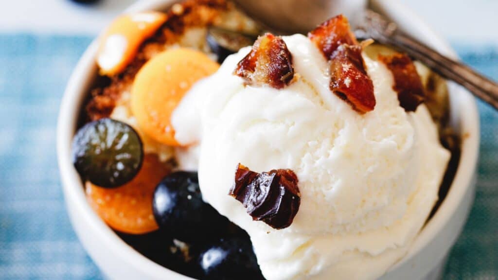 A bowl of dessert featuring a scoop of vanilla ice cream, sliced fruit, and chopped nuts on top, with a spoon at the side.