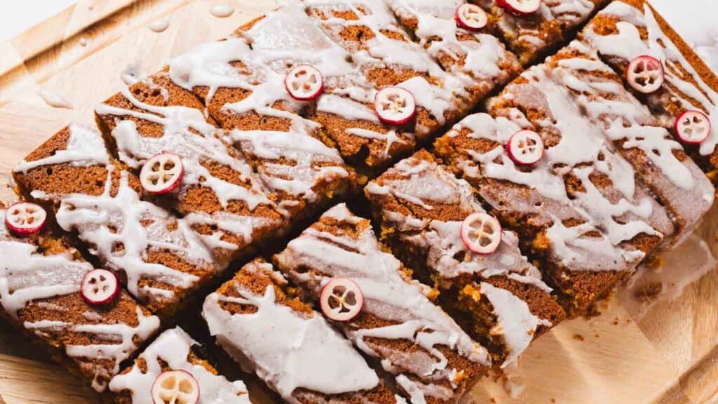 Sliced loaf cake with white icing drizzle and small red fruit slices on a wooden board.