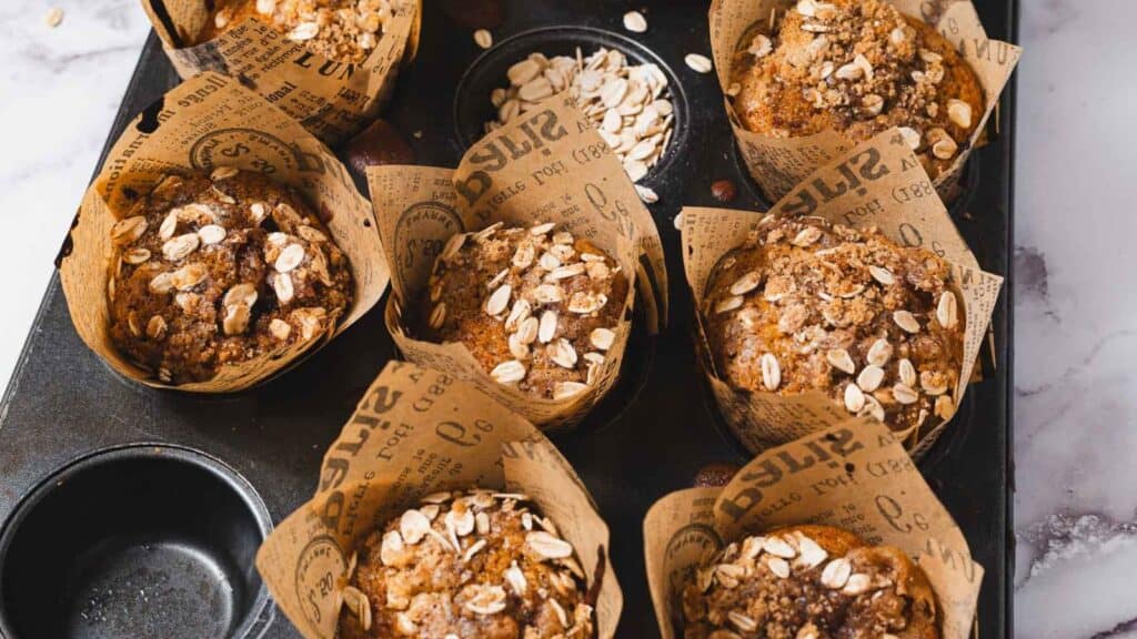 Seven muffins with oat toppings in paper liners on a baking tray, with some oats scattered in an empty tray section.