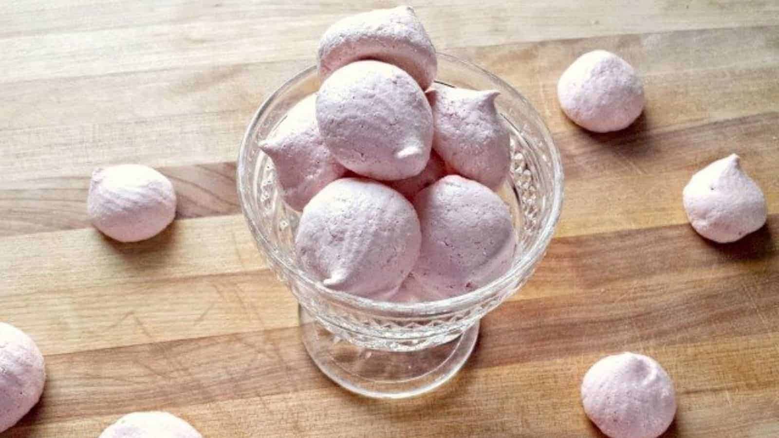 Image shows Raspberry Meringues in a glass parfait dish with more surrounding it on a wooden board.