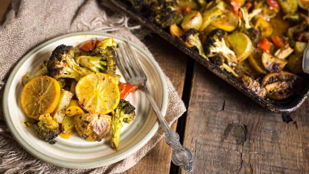 A plate of roasted vegetables, including broccoli, lemons, and potatoes, with a fork on a wooden table. A baking tray with more vegetables is in the background.