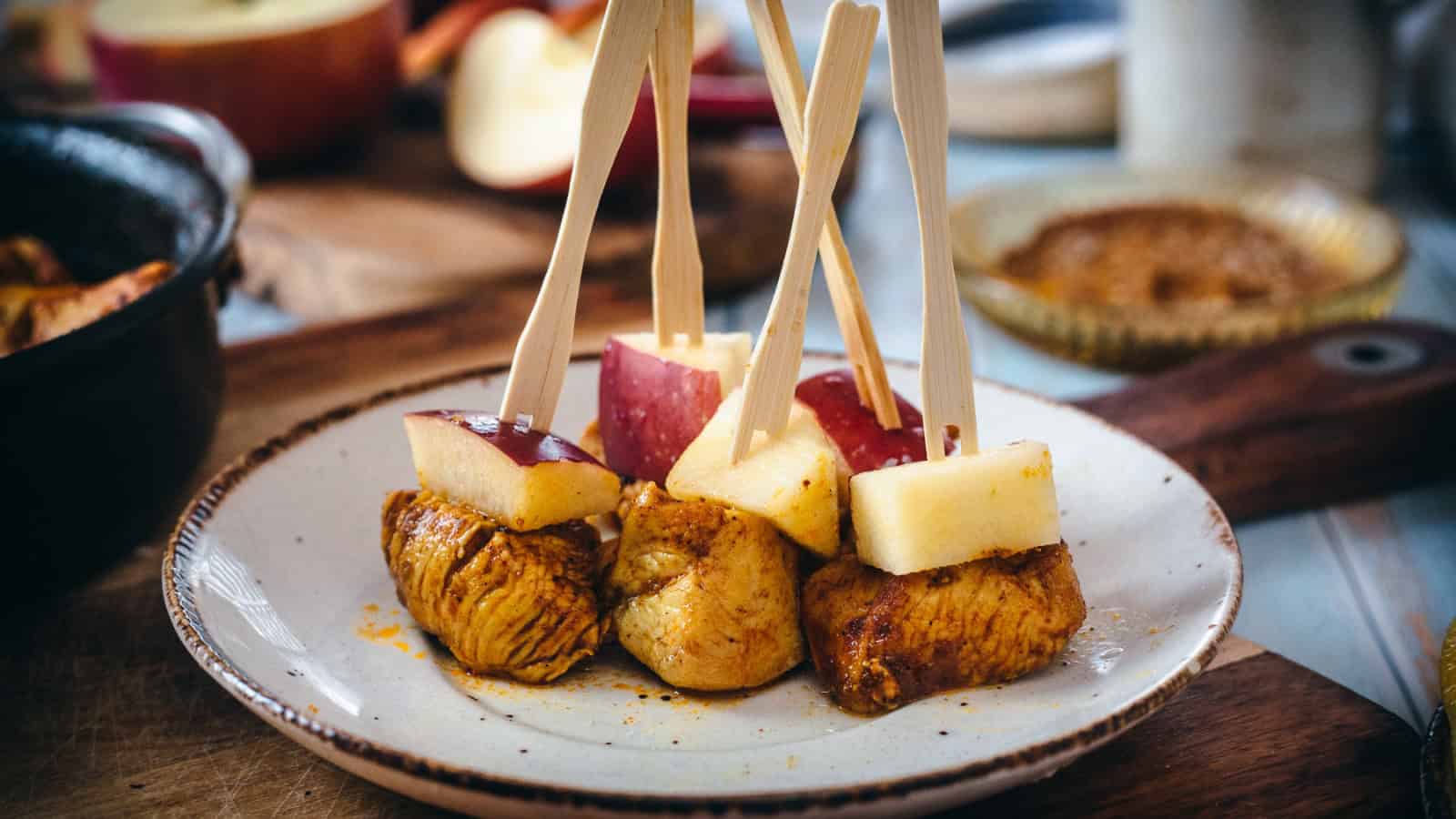 A plate contains skewers with cooked meat cubes and red apple pieces. The skewers are upright, resting on the plate. In the background, there are additional food items and a blurred bowl with a brown dip.