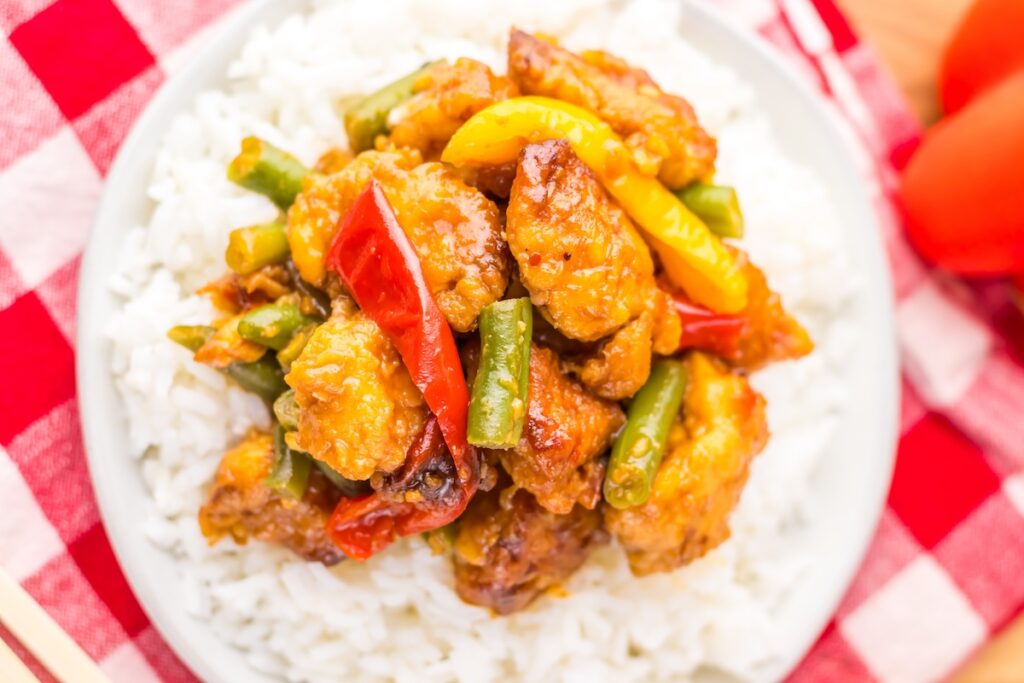 A plate of stir-fried chicken with vegetables, including red and yellow peppers and green beans, served over a bed of white rice on a checkered tablecloth.