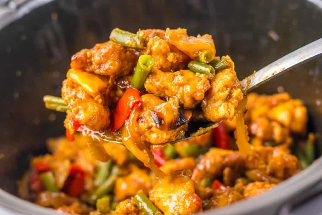 Close-up of a spoon lifting a portion of stir-fried chicken with green beans, red bell peppers, and onions in a flavorful sauce from a black pot.